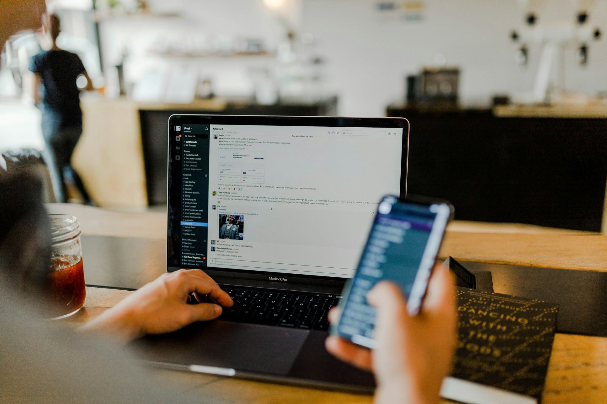 Hands on a laptop keyboard while holding a phone at a cafe table